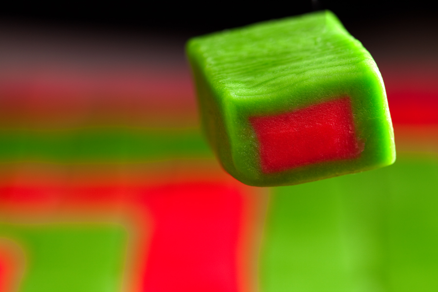 This is a Brian Charles Steel photograph of a piece of red and green Hubba Bubba gum floating above red and green gum.  The piece is rectangular; it is red in the center and green on the outside. 